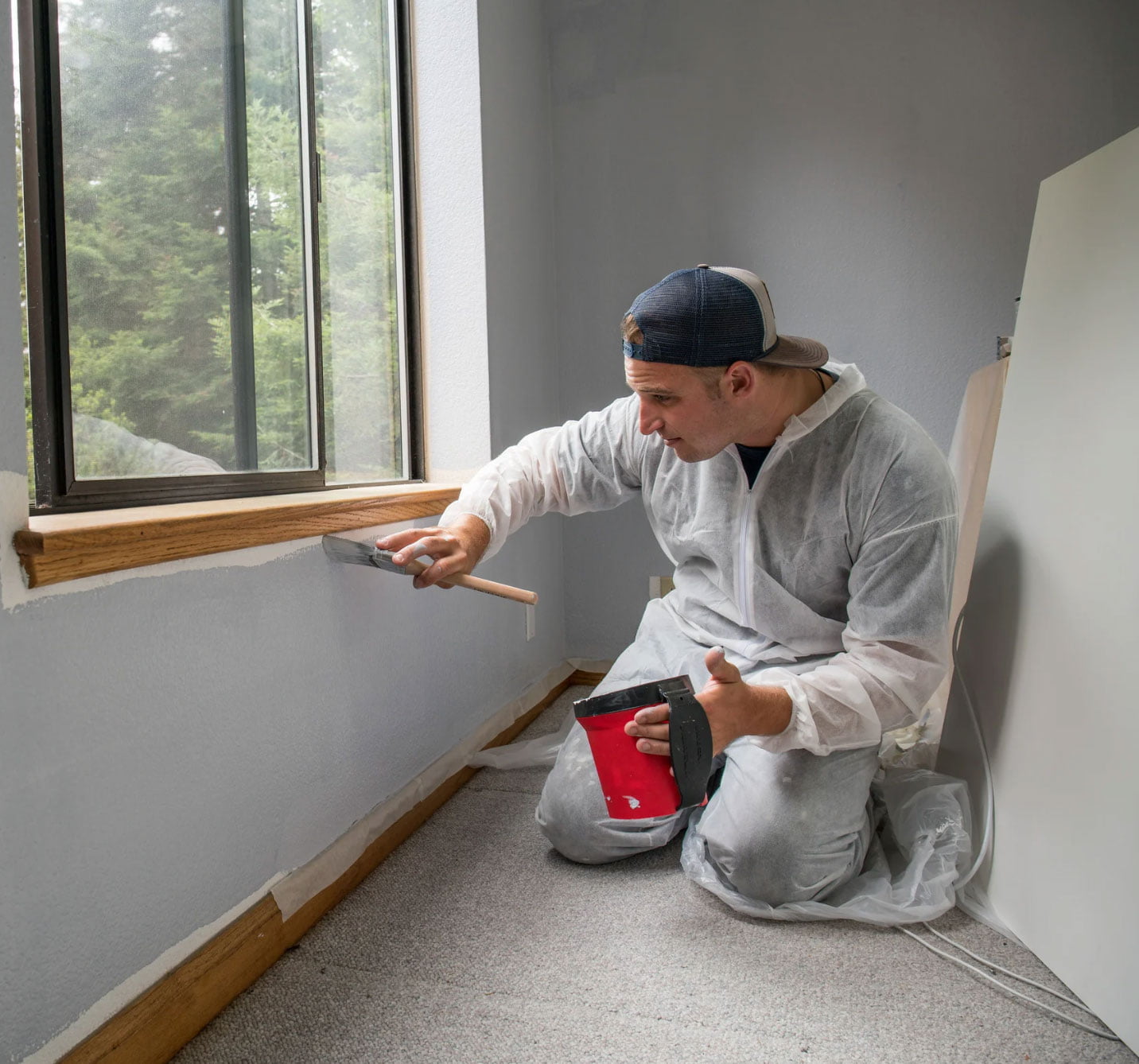 Person wearing overalls painting a window sill in a room with gray walls and a view of trees outside.