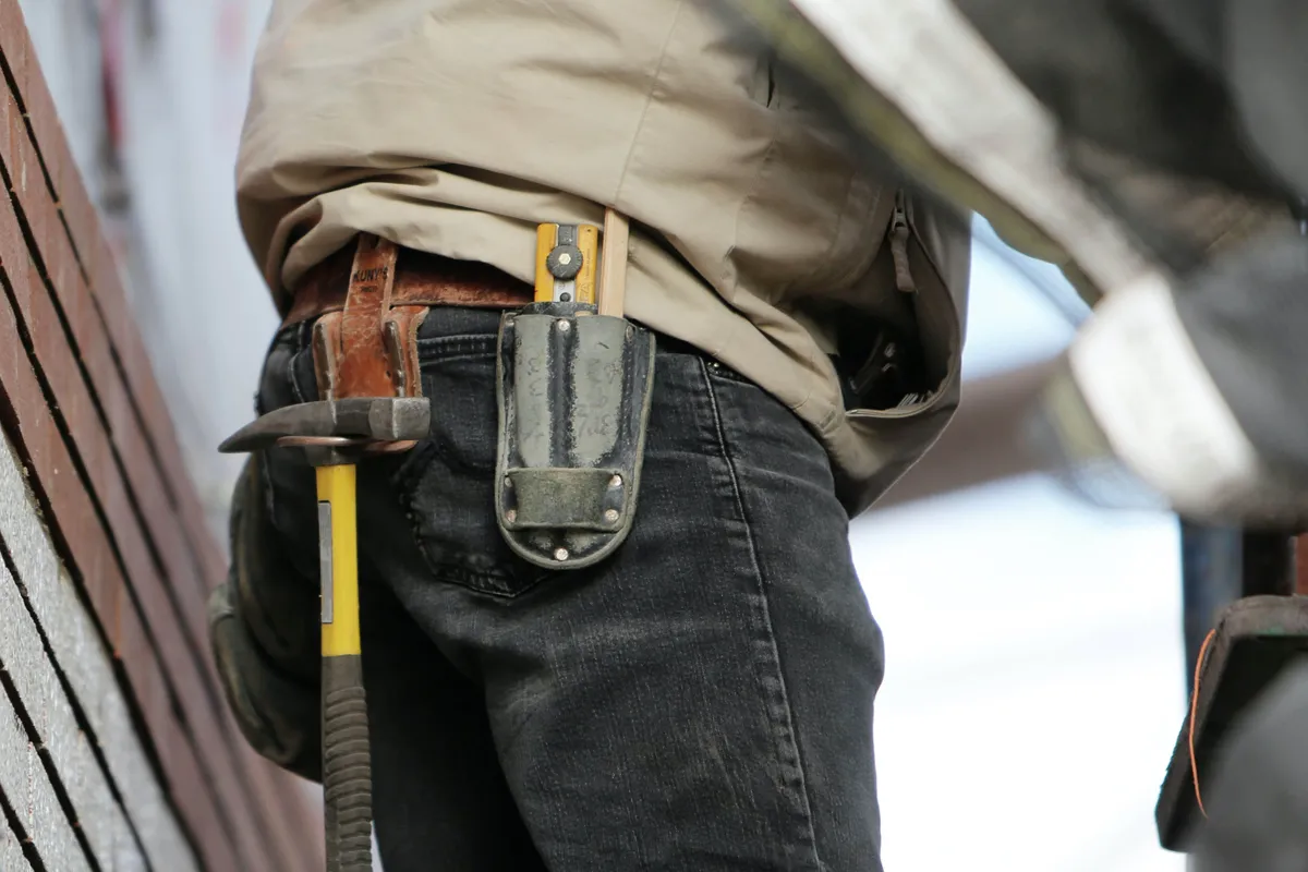 benefits 1 A construction worker with tools in their belt, wearing jeans and a jacket, stands next to a brick wall.
