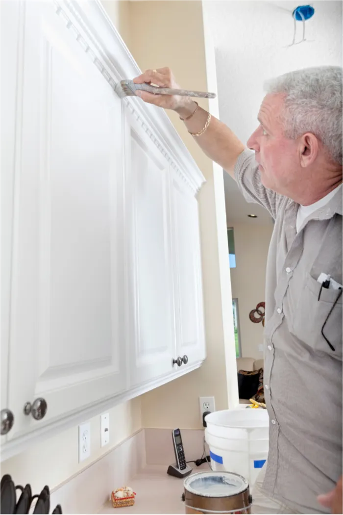 Person painting white kitchen cabinets above a countertop with tools and supplies nearby.