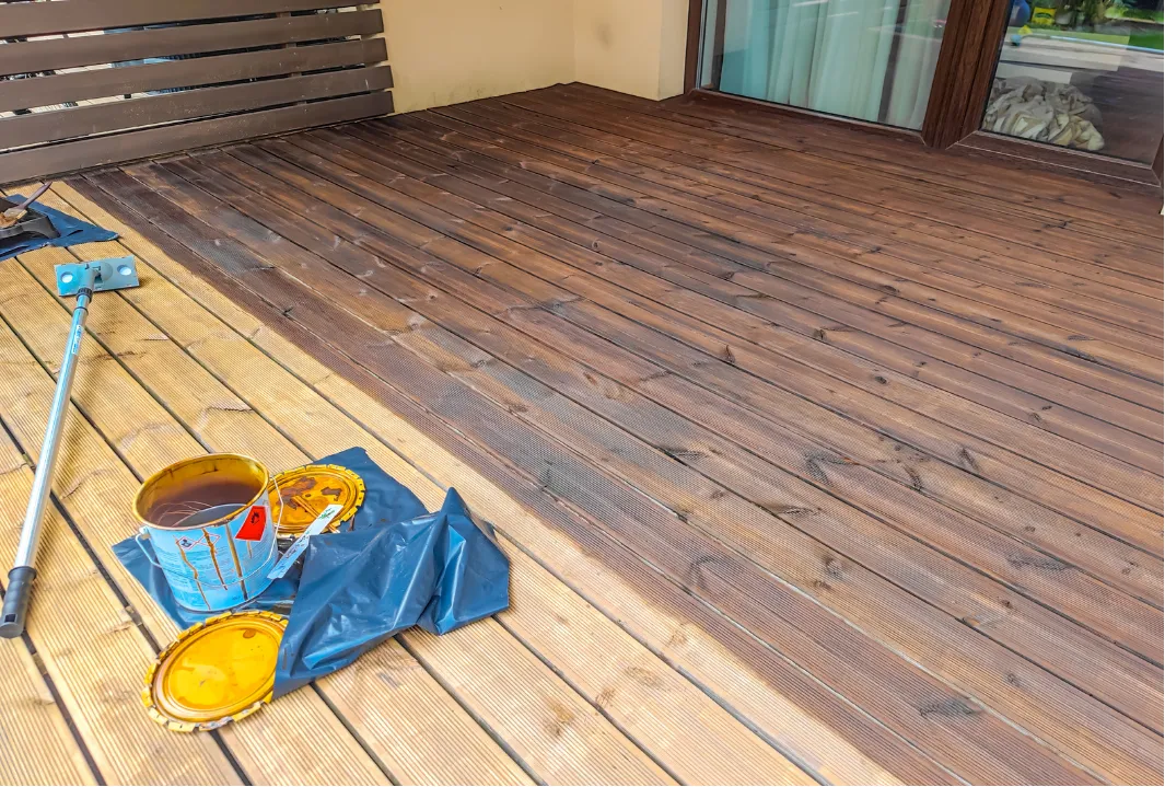 Fence Painting, All in House LLC in Tualatin, OR: A wooden deck half-stained, with open wood stain cans and a roller brush lying nearby.