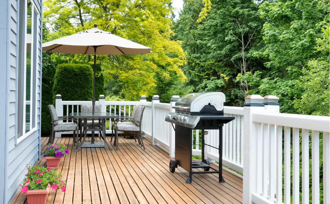 deck-and-patio-construction-2 Patio Construction - All In House LLC in Tualatin, OR: Wooden deck with a grill, round table, umbrella, chairs, and potted flowers, surrounded by lush green trees.