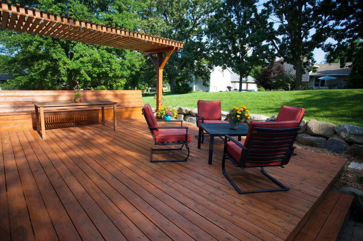deck-and-patio-construction-3 Wooden patio with red cushioned chairs, a table, and a pergola. Lawn and trees in the background on a sunny day.