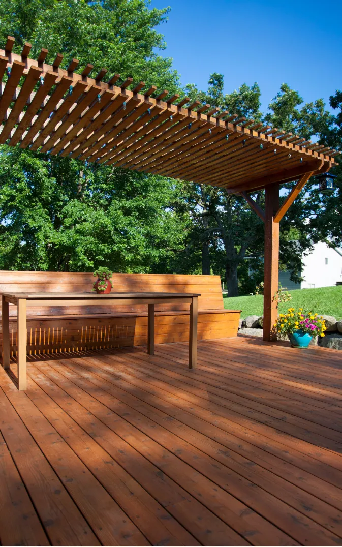 deck-and-patio-construction-4 (1) Wooden deck with a pergola, a bench, and a table. A flower pot sits on the table, with trees and lawn in the background.