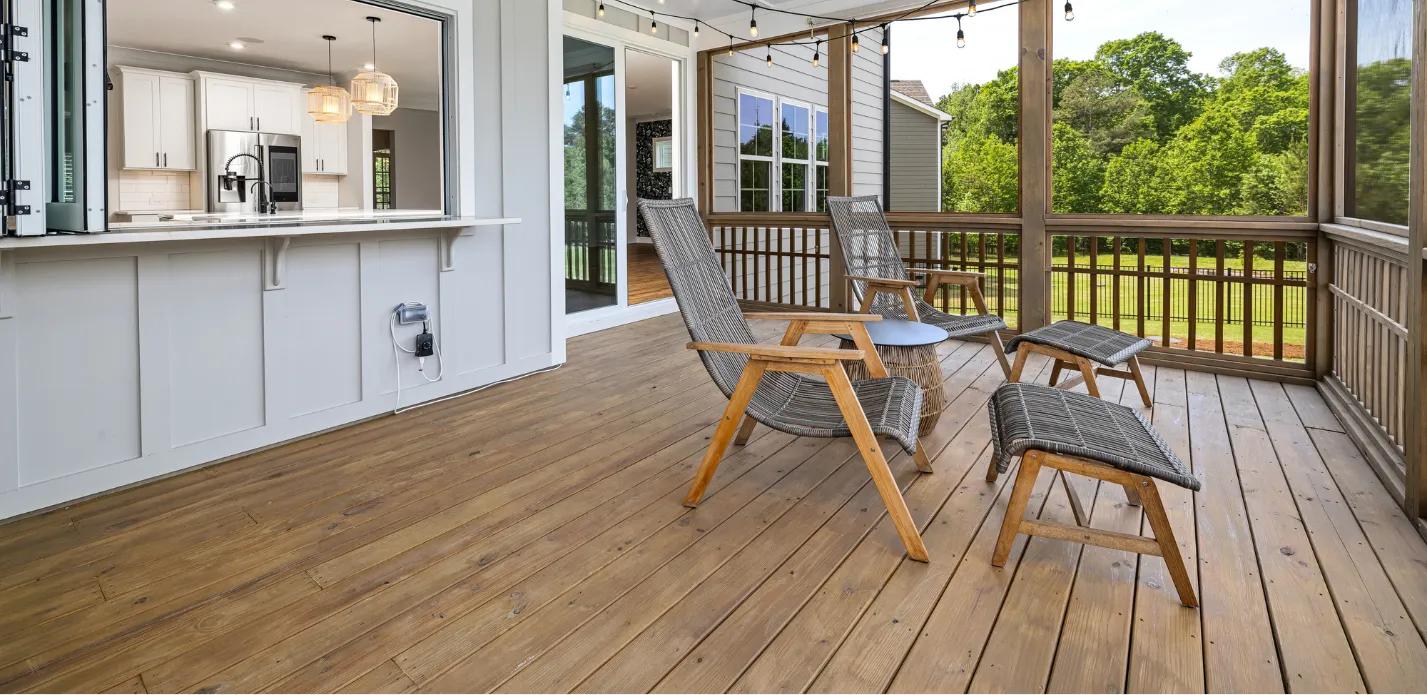 deck-and-patio-construction-c Spacious wooden deck with chairs, a small table, and a view of greenery. Open kitchen window in the background.