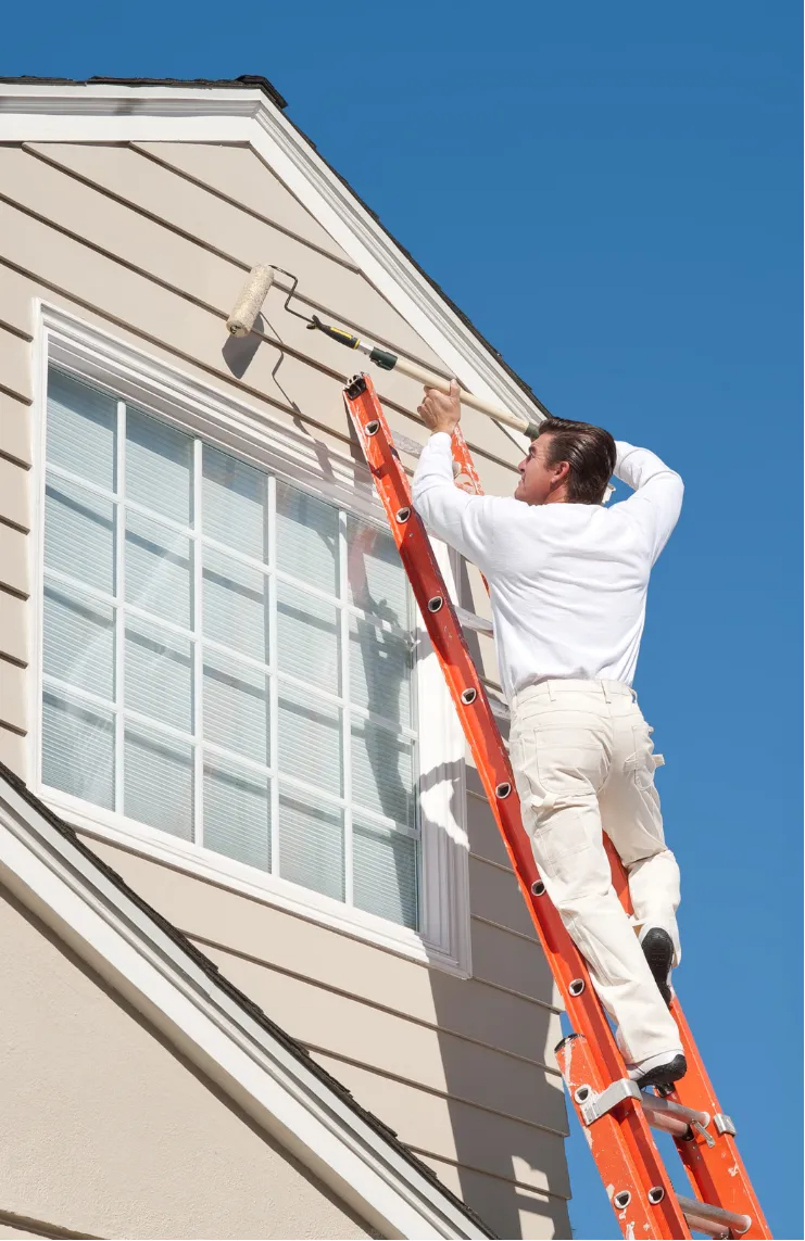 Person on a ladder painting the exterior of a house near a window against a clear blue sky.