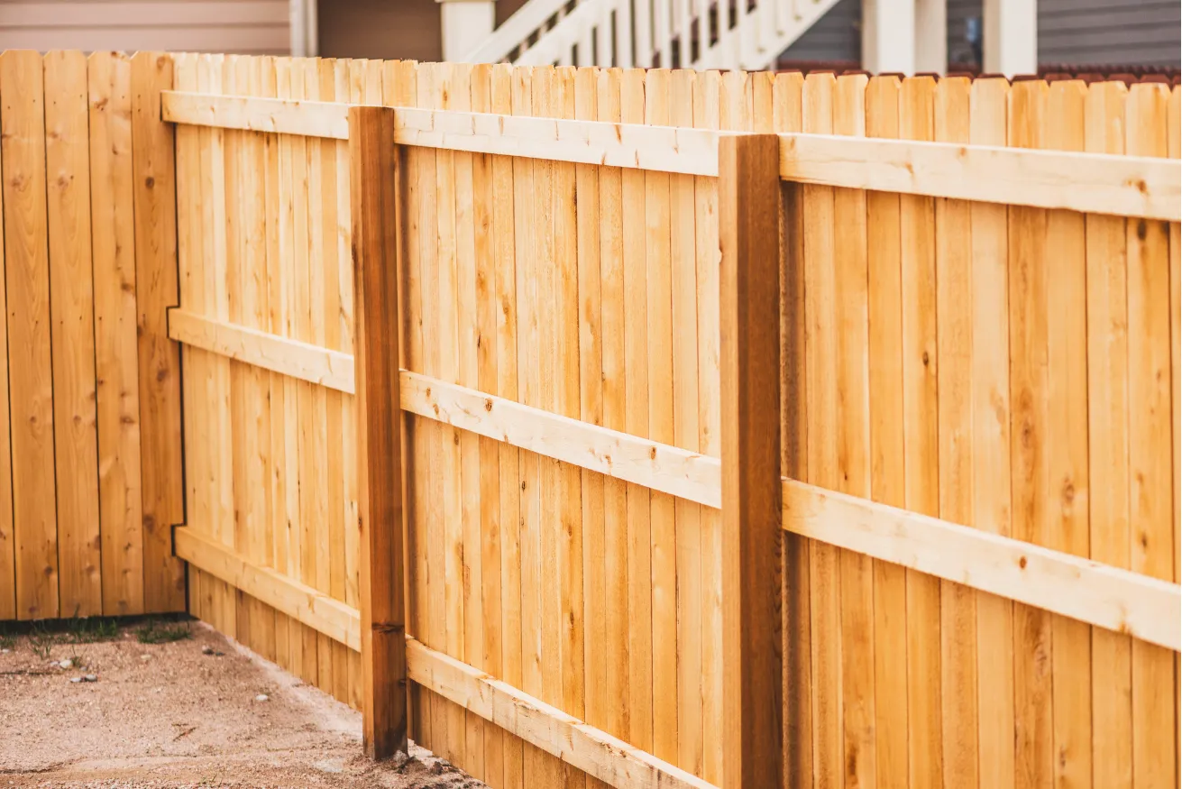 fence-install-1 Wooden fence with vertical planks, supported by horizontal beams, surrounding a yard with houses in the background.
