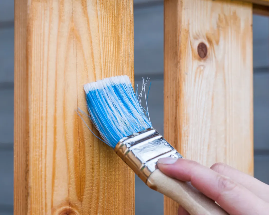 Close-up of a hand painting wooden planks with a paintbrush, applying a blue hue to a natural wood surface.