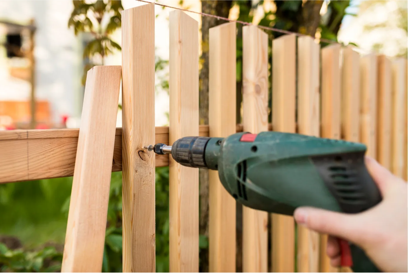fence-repair-1 Close-up of a person using a green power drill to fasten wooden planks on an outdoor fence.