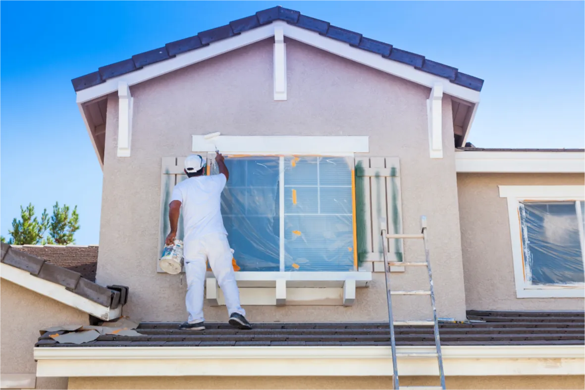 A painter in white overalls paints the exterior of a house while standing on the roof beside a ladder.