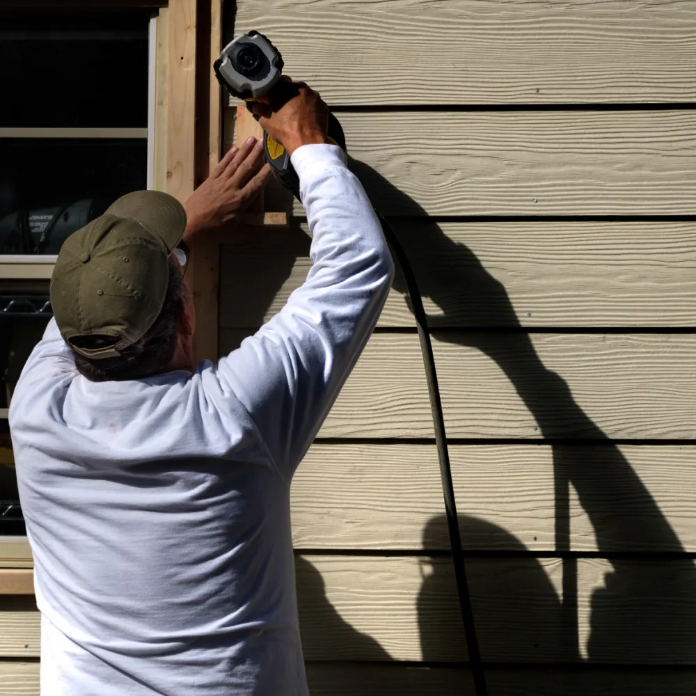 siding-install Person in white shirt and cap using a power tool on exterior siding, casting a shadow on the wall.