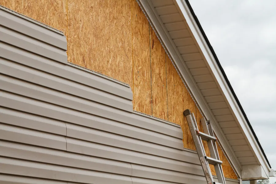 Partially installed beige vinyl siding on a house exterior with a ladder leaning against it.