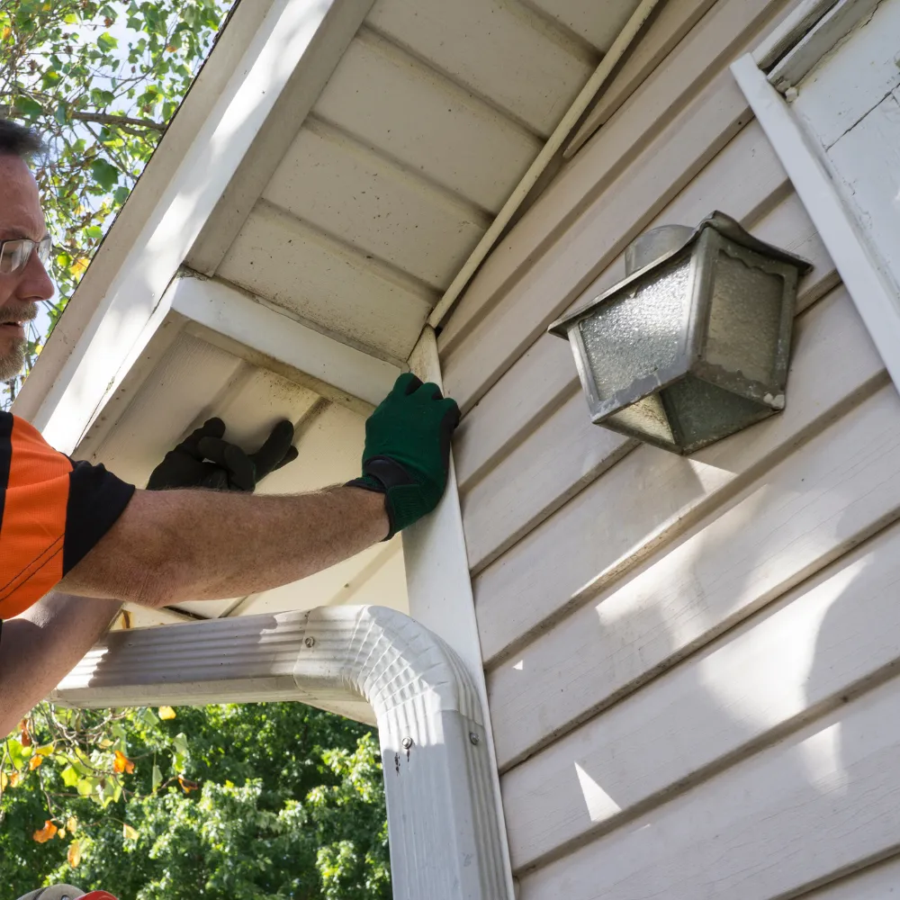 Person in gloves installing siding on a house using a ladder, with a nearby outdoor light fixture.