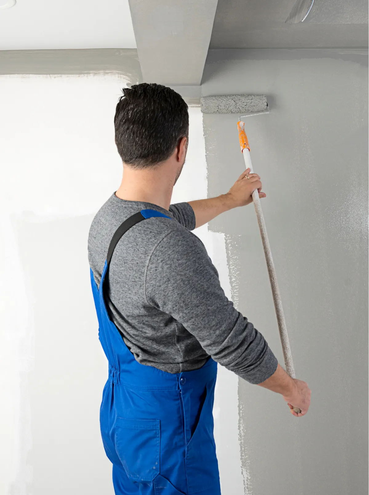 Person in blue overalls and gray shirt painting a wall gray with a roller on an extension handle.