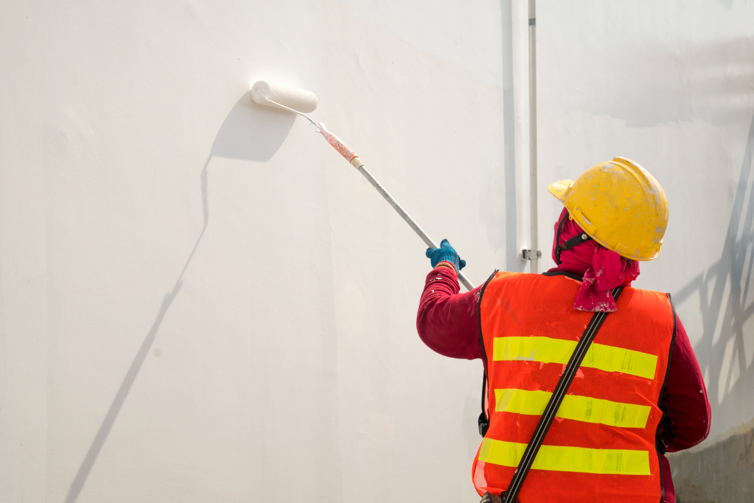 570 Worker in a safety vest and helmet paints a white wall using a roller on an extendable pole.