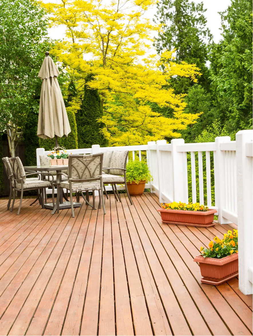 deck-1 Wooden deck with patio table, chairs, umbrella, potted flowers, and green trees in the background.