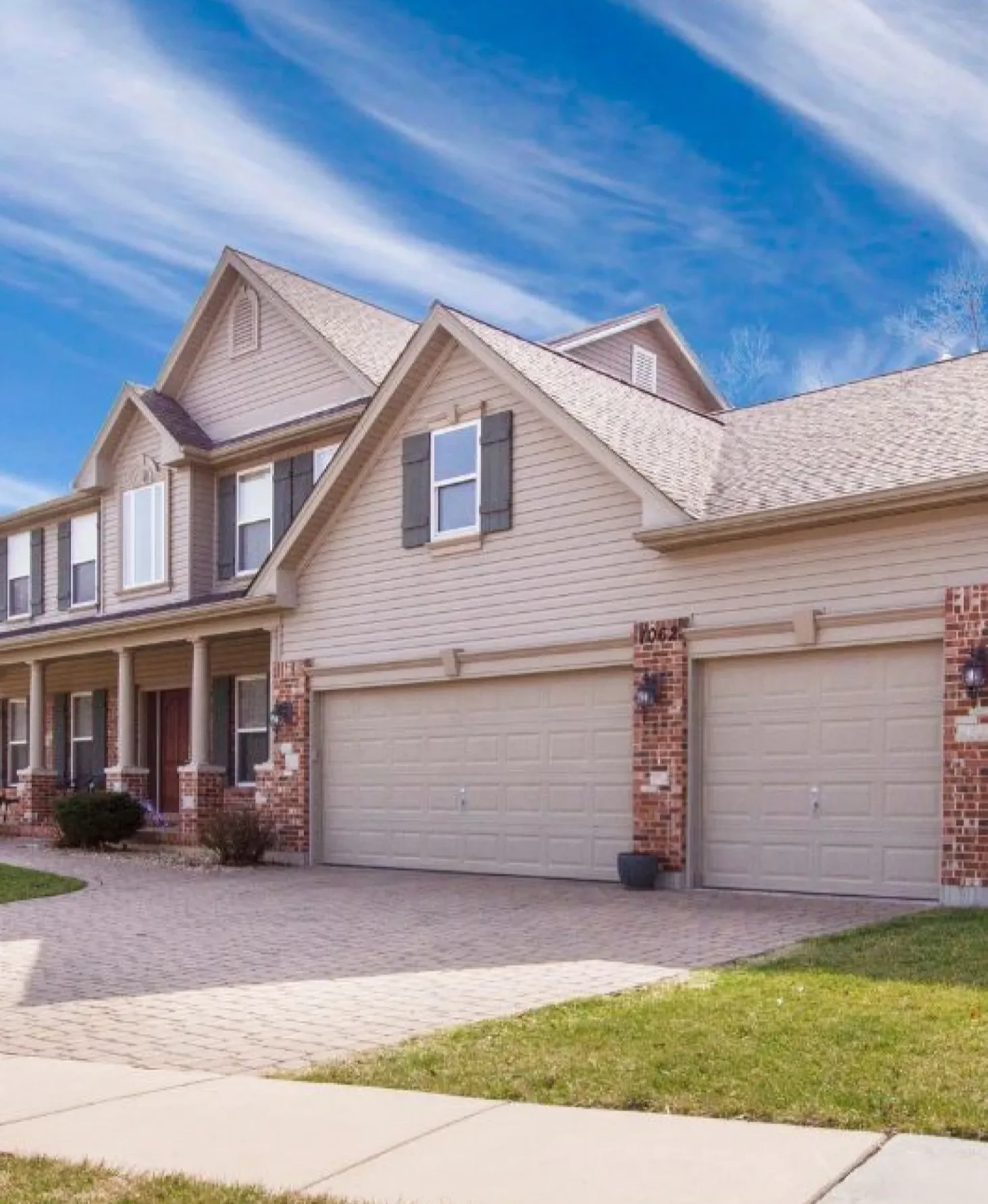 Two-story suburban house with a three-car garage, brick accents, and a bright blue sky above.