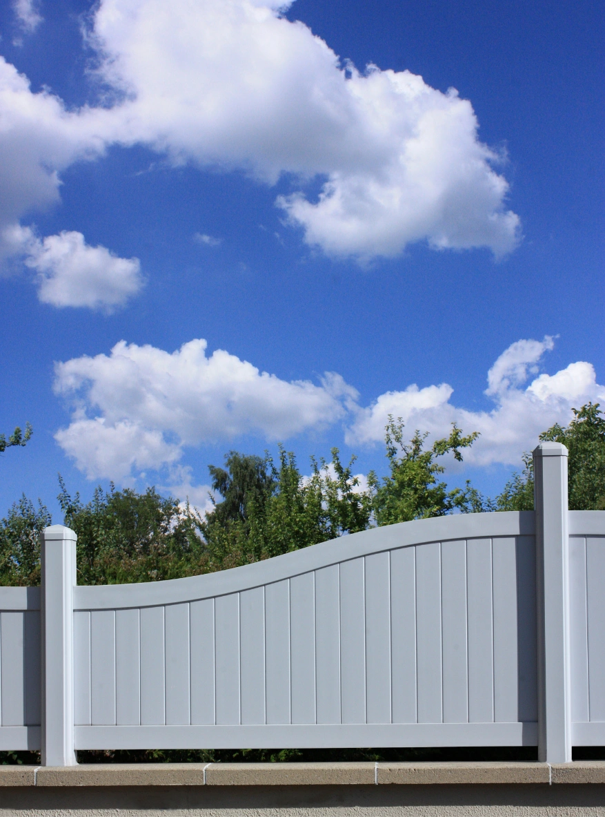fence-1 White vinyl fence with a wavy top, green trees behind it, and a blue sky with scattered clouds above.