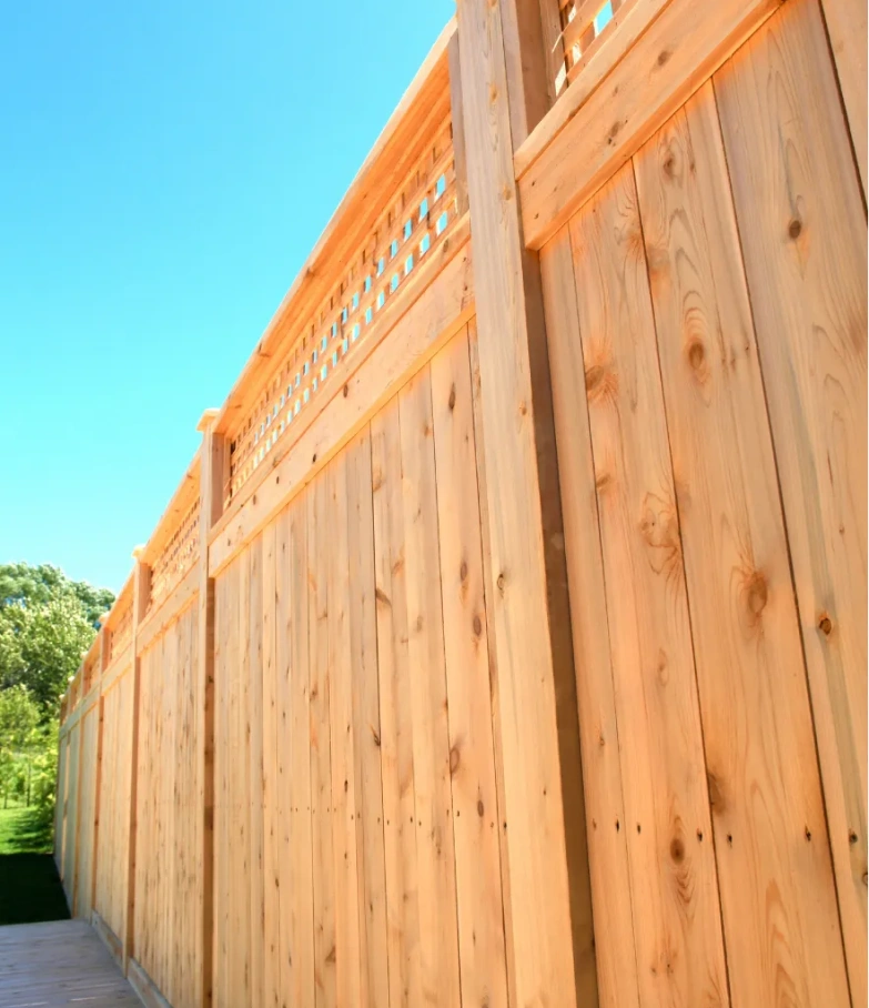 Tall wooden privacy fence with decorative lattice on top, set against a clear blue sky and greenery.