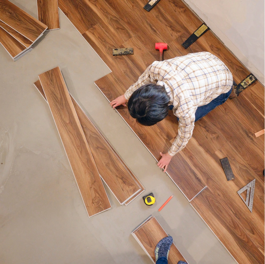 floor-replace-2 Person installing wooden floor panels, surrounded by tools and measuring tape on a partially finished floor.