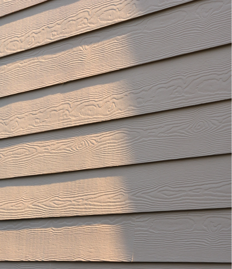 siding-rep-2 Close-up of beige house siding with wood grain texture, partially lit by sunlight.