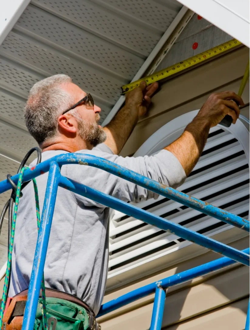 siding-w A man on a blue scaffold measures siding under a roof eave with a tape measure on a sunny day.