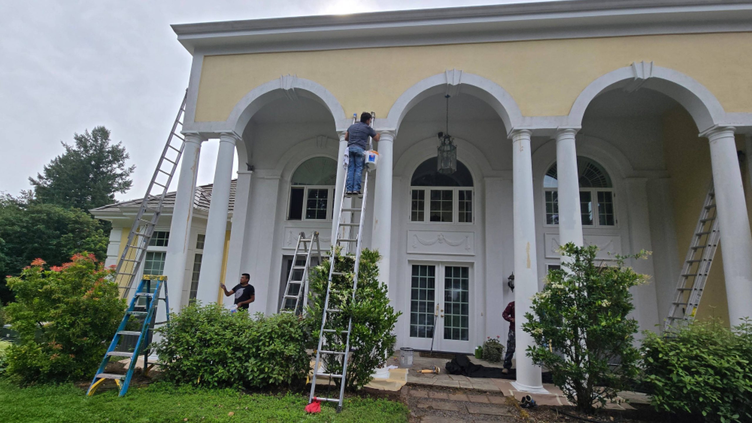 Workers on ladders paint the exterior of a two-story white and yellow house with arched porticos.