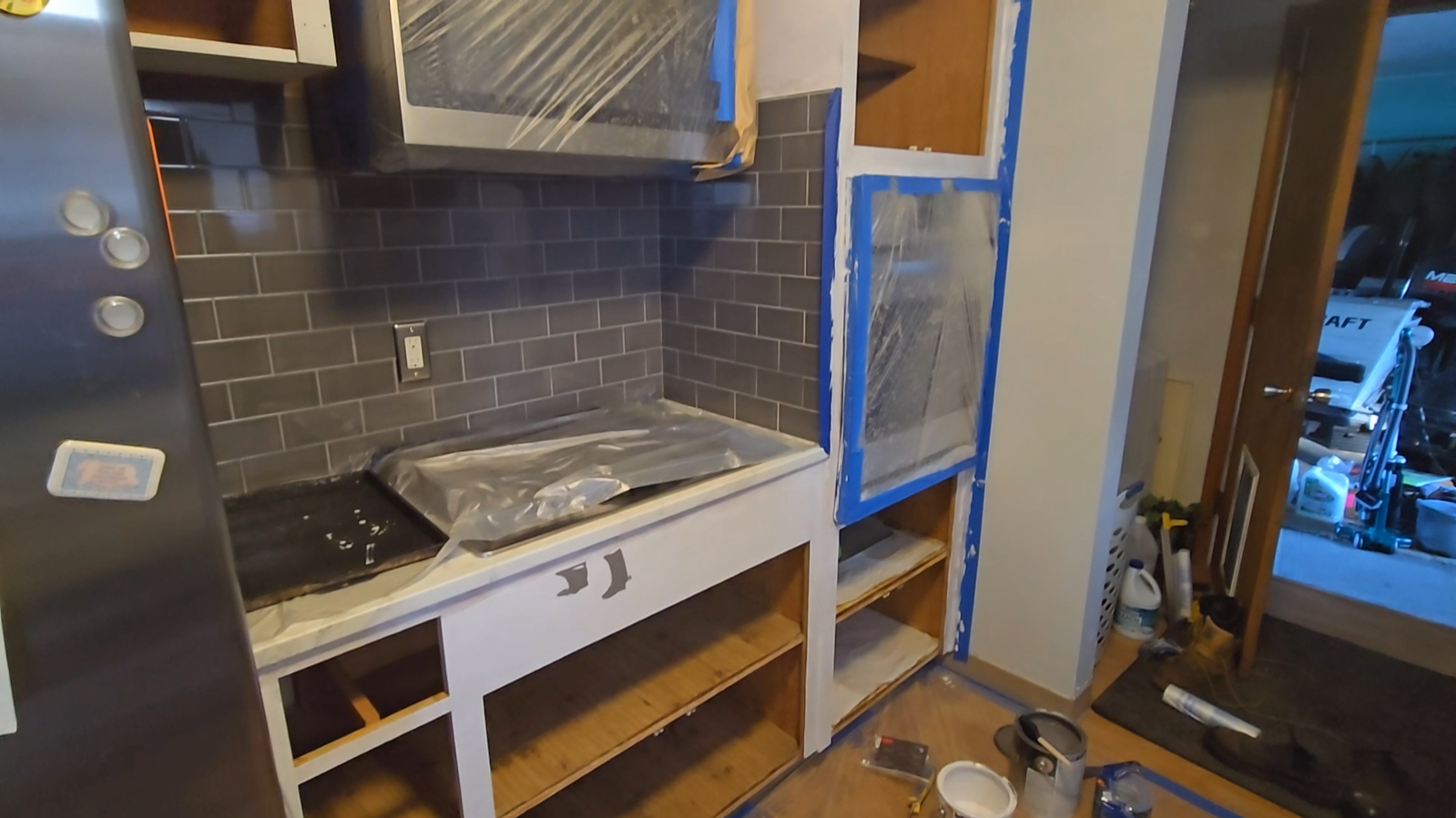 Kitchen renovation in progress with bare cabinet frames, blue painter’s tape, and gray subway tile backsplash.