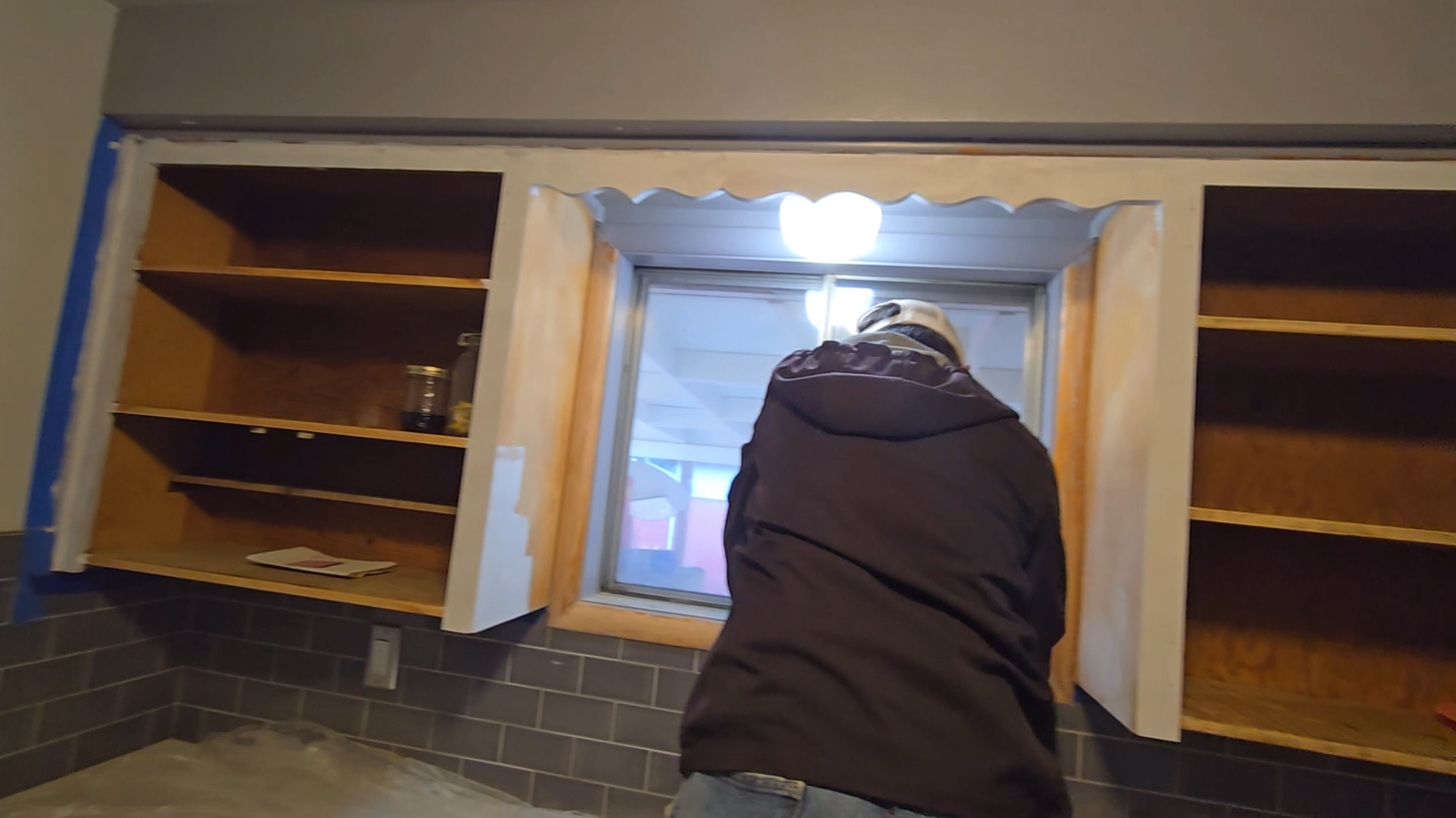 A person in a hooded jacket working on window trim between two open kitchen cabinets.