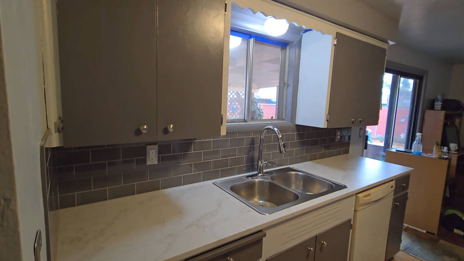 Kitchen sink and counter with grey cabinets and a tiled backsplash.